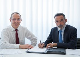 Two businessmen discussing and signing documents at a bright office table.